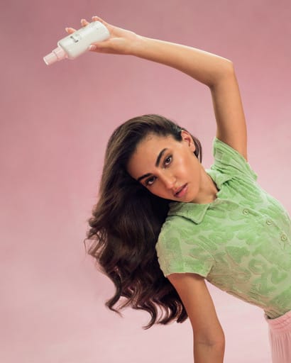 A product frame on a pink ground: a model in a green top holding a white bottle overhead.