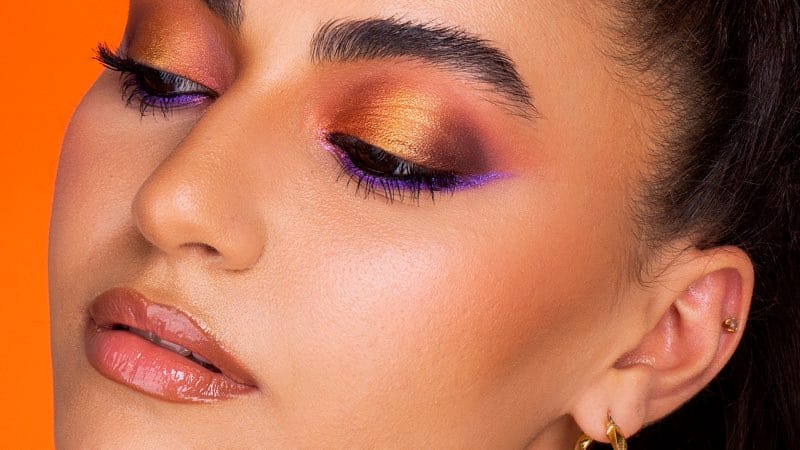 A beauty frame on an orange ground: warm copper and plum eyeshadow with a violet lower line.