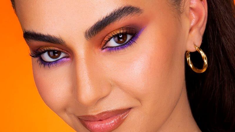 A beauty frame on an orange ground: gold and copper shadow with a violet graphic line, eyes to camera.