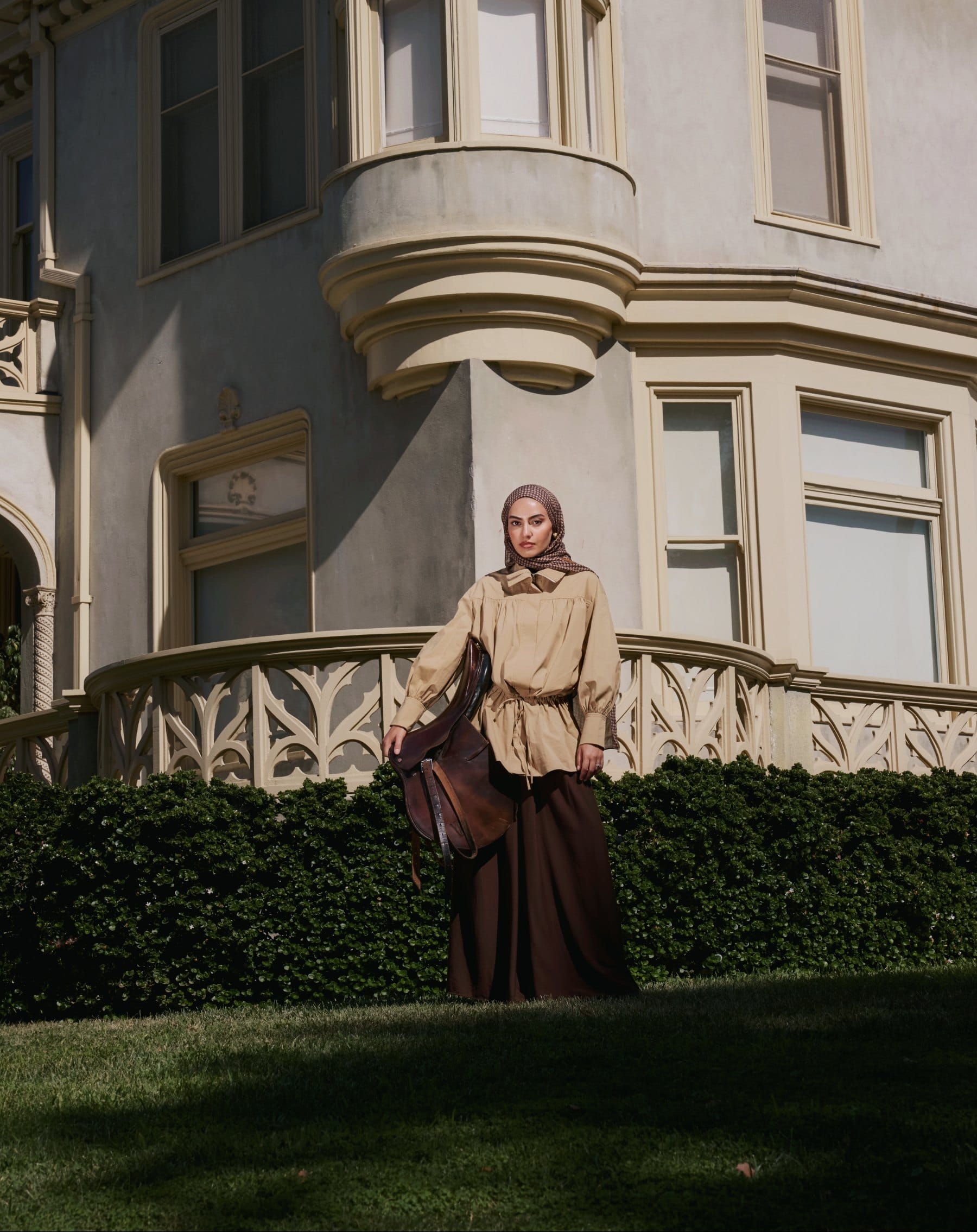 Campaign photograph: a model in a houndstooth headscarf and tan blouson standing on a lawn outside a Gothic-revival manor, holding a leather saddle.